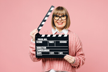 Elderly woman 50s years old wear sweater shirt casual clothes glasses hold in hand classic black film making clapperboard isolated on plain pastel pink background studio portrait. Lifestyle concept.