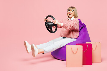 Full body elderly young woman wears casual clothes hold package bags after shopping sit in bag chair, steering wheel driving car isolated on plain pink background. Black Friday sale buy day concept.