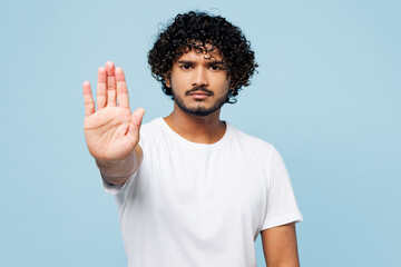 Young serious strict sad Indian man wear white t-shirt casual clothes showing stop gesture with palm look camera isolated on plain pastel light blue cyan background studio portrait. Lifestyle concept.
