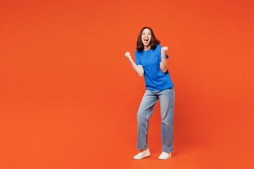 Full body young smiling surprised excited fun happy woman she wear blue t-shirt casual clothes do winner gesture clench fist isolated on plain red orange background studio portrait. Lifestyle concept.