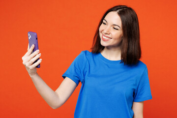 Young smiling happy woman she wears blue t-shirt casual clothes doing selfie shot on mobile cell phone post photo on social network isolated on plain red orange background studio. Lifestyle concept.