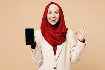 Young Arabian Asian Muslim woman wear red abaya hijab suit clothes use blank screen area mobile cell phone show ok okay isolated on plain beige background. UAE middle eastern Islam religious concept.