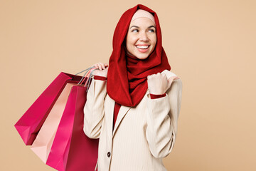 Young Arabian Asian Muslim woman wears red abaya hijab suit clothes shopping hold package bags point finger aside isolated on plain beige background. Black Friday sale day UAE Islam religious concept