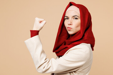 Side view young Arabian Asian Muslim woman wear red abaya hijab suit clothes show hand biceps muscles demonstrating power isolated on plain beige background UAE middle eastern Islam religious concept
