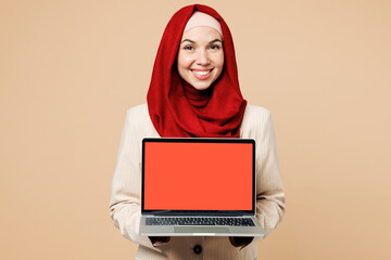 Young Arabian Asian Muslim IT woman wear red abaya hijab suit clothes hold use work on blank screen laptop pc computer isolated on plain beige background. UAE middle eastern Islam religious concept.