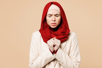 Young sad Arabian Asian Muslim woman she wearing red abaya hijab suit clothes hold hands folded in prayer gesture begging isolated on plain beige background UAE middle eastern Islam religious concept.