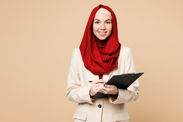 Young Arabian Asian Muslim woman wears red abaya hijab suit clothes hold clipboard with paper account documents isolated on plain beige background studio. UAE middle eastern Islam religious concept.