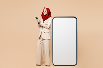 Young Arabian Asian Muslim woman wear red abaya hijab suit clothes stand big huge blank screen free area mobile cell phone use smartphone device isolated on plain beige background. UAE Islam concept.