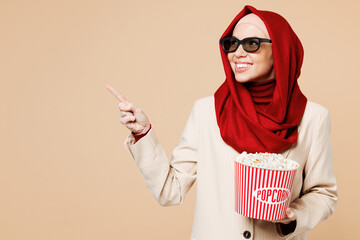 Young Arabian Asian Muslim woman wear red abaya hijab suit clothes 3d glasses watch movie film hold bucket of popcorn in cinema point aside look aside isolated on plain beige background. UAE concept.
