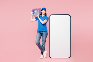 Full body happy delivery employee woman wears blue cap t-shirt uniform workwear work as dealer courier big huge blank screen area mobile cell phone hold water bottle isolated on plain pink background.