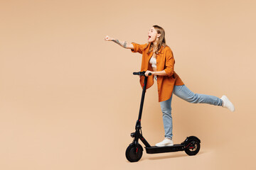 Full body side view young woman wear orange shirt casual clothes ride electric scooter do super hero power gesture isolated on plain pastel light beige background studio portrait. Lifestyle concept.