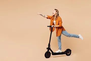 Tuinposter Full body side view young woman wear orange shirt casual clothes ride electric scooter do super hero power gesture isolated on plain pastel light beige background studio portrait. Lifestyle concept. © ViDi Studio