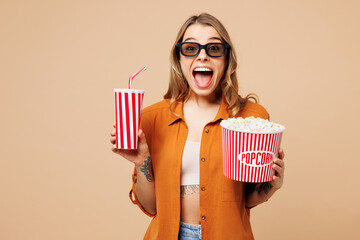 Young shocked fun woman wear orange shirt casual clothes 3d glasses watch movie film hold bucket of popcorn cup of soda pop in cinema isolated on plain pastel light beige background studio portrait. - 791453924
