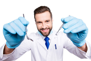 Portrait of positive comic dentist with stubble in white lab coat, blue tie in working process on grey background, treat, examine cavity, searching for sick tooth, holding tools in hands