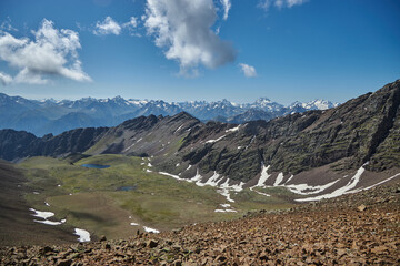 Panoramic vista from a mountain summit showcasing a range of snow-dusted peaks, a clear blue sky overhead, and a serene alpine lake