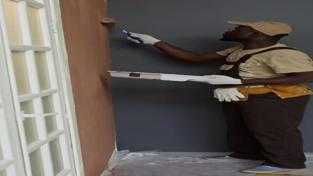 Vertical side full footage of Black male handyman painting walls with brown color using paint roller while working and redecorating new apartmentVertical side full footage of Black male handyman paint