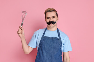 Portrait of happy confectioner with funny artificial moustache holding whisk on pink background