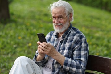 Portrait of happy grandpa with glasses using smartphone on bench in park