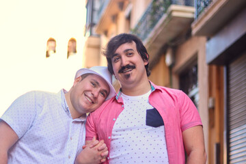Couple of gay men arm in arm smiling walking down the street, one rests his head on the other,...
