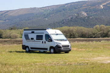 A modern white camper van is parked on a grassy field with hills and wind turbines in the...