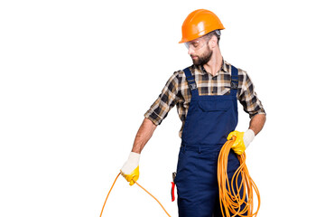 Half face portrait of busy stylish electrician with stubble in overall, shirt, hardhat installing, laying a cable, having rolled wire in hand, standing over grey background