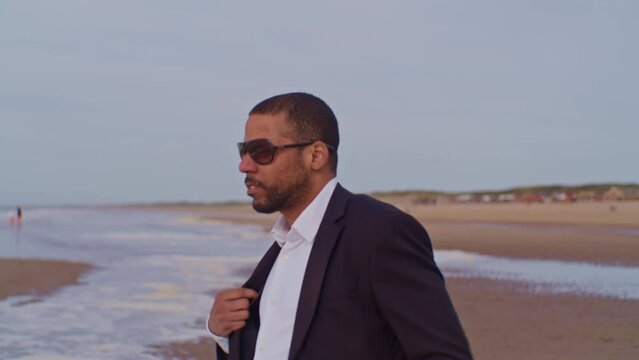 A male man black latino model in a suit walks on the beach and takes off his jacket at the seashore with sunglasses in the Netherlands, the Hague