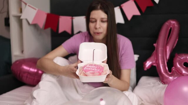 Young millinial brunette woman pink t-shirt making wishes and blowing out candles delicious pink bento cake with inscription happy birthday white box bedroom on bed decorated with flags and balloons