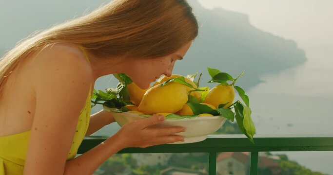 A woman smells fresh lemons on a balcony overlooking the sea. The image evokes the senses and the beauty of a serene landscape.