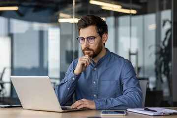Serious young business man works in the office with a laptop, sits at the desk and looks...