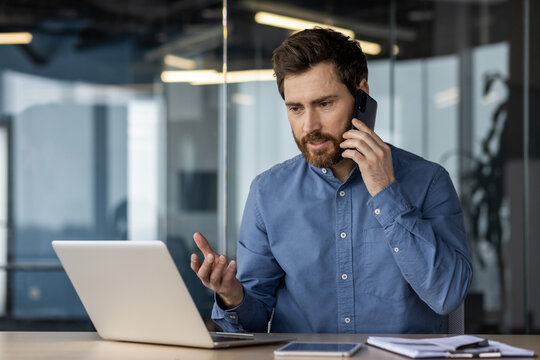 Young serious business man sitting in the office at the desk, talking on the mobile phone, explaining and discussing, pointing at the laptop screen