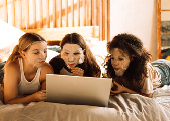 Charming mixed-race teenagers in cosmetic fabric masks on their faces are looking at laptop....