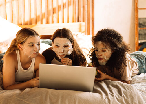 Charming mixed-race teenagers in cosmetic fabric masks on their faces are looking at laptop. Multiethnic friends have fun and use computer. - Powered by Adobe