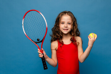 Cheerful little sports girl in red suit playing tennis isolated over blue background. Sport, study,...