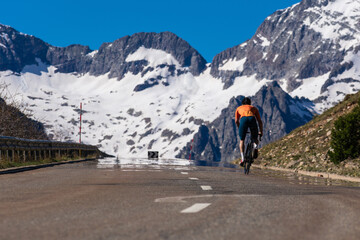 A man rides a bicycle. He is riding away on the road with snow-capped mountains in the background.