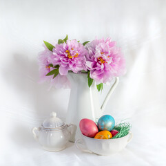 Easter still life with gorgeous pink peonies in a vase and coloured easter eggs in a fine porcelain bowl on a white background.
