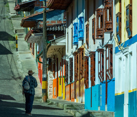 Male, farmer, walking in the street with colored facades in Jerico, Jericó, Antioquia, Colombia. Wooden carved balconies. The man wears a wooden mocjila and a typical hat. Shadows.