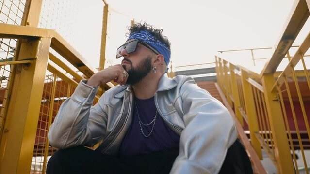 A stylish bearded man wearing a headband and sunglasses poses thoughtfully in an urban environment, exuding an air of cool confidence.