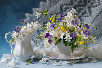 Beautiful still life with spring flowers, a bouquet of white and blue flowers in a cup and jug on a decorative background with a white shawl. - 791536124