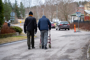two men walking on a street. the other man using a rollator
