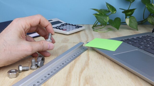 Man's hand holds a screw, on desk in engineering office with compiuter and ruler.