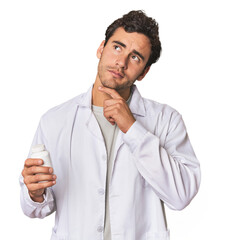 Young Hispanic pharmacist with pills looking sideways with doubtful and skeptical expression.