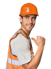 Young Hispanic worker in safety gear points with thumb finger away, laughing and carefree.