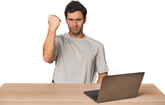 Hispanic young man working on laptop showing fist to camera, aggressive facial expression.