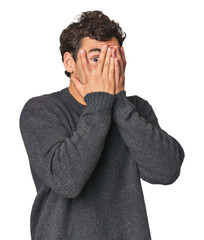 Young Hispanic man in studio blink through fingers frightened and nervous.