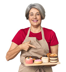 Caucasian mid-age baker with pastry tray smiling and pointing aside, showing something at blank space.