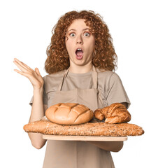 Young Caucasian redhead woman holding bread tray surprised and shocked.