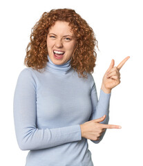 Young Caucasian redhead woman pointing with forefingers to a copy space, expressing excitement and...