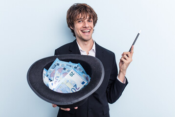 Young magician man holding a hat with banknotes isolated on blue background