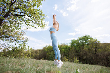 The girl has her hands up and is standing on her toes thus she is stretching in the park. Beautiful blonde Caucasian woman in blue tight tracksuit. Blonde girl at an outdoor training session