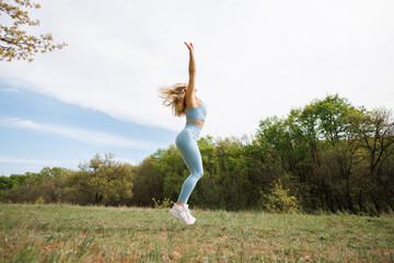 The girl performs jumps with her hands in the air. Beautiful blonde Caucasian woman in blue tight tracksuit. Blonde girl at an outdoor training session